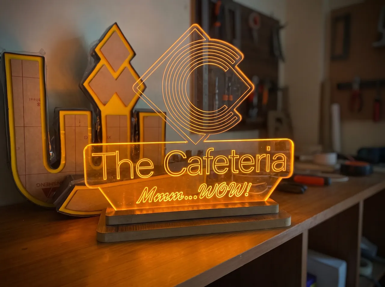 Edge-lit clear acrylic sign glowing amber with The Cafeteria logo, on a workshop bench.