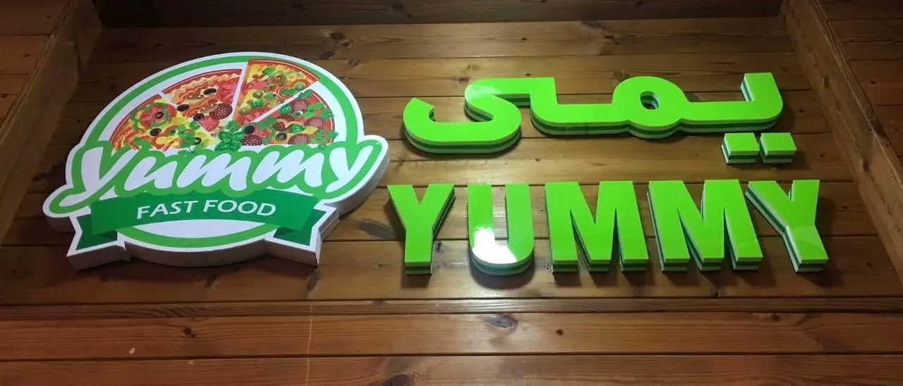 Yummy Fast Food sign: green layered acrylic letters in Dari and English beside a round pizza logo, on timber.